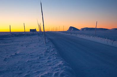 Eine Straße zieht sich durch Schneelandschaft zu Beginn der Blauen Stunde, während das Restlicht der Sonne den Horizont gelb-orange färbt.