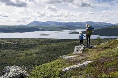 Kvinne og barn står på liten stein og skuer utover et storslått landskap, med innsjø og fjell i bakgrunnen.