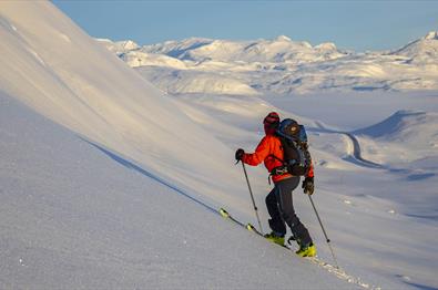 Person på vei opp fjellside med randoneeski, vid utsikt over snødekt fjellandskap