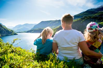 En familie sitter i en li og nyter utsikt over vann og fjell.