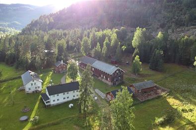 Dronenfoto des Hofes mit Gebäuden, großen Rasenflächen und angrenzendem Birkenwald. Sonnenstrahlen im gegenlicht erzeugen ein Mosaik aus Licht und Schatten.