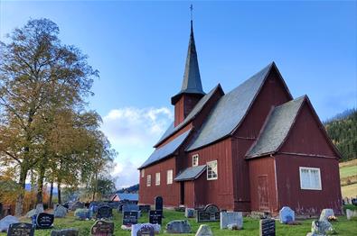 A red-brown stave church surrounded by a church yard with birch trees that start to get autumn-coloured leaves.