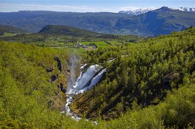 En dobbel foss som spruter mye vann. Bygd og fjell i bakgrunnen.