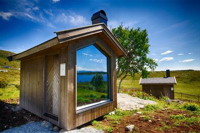 En bitte liten moderne trehytte med stort utsiktsvindu på et grønt stølsjorde på fjellet.