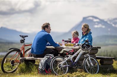 A family on a bicycle trip takes a break at a rest area with picknick table. Mountains in the background.