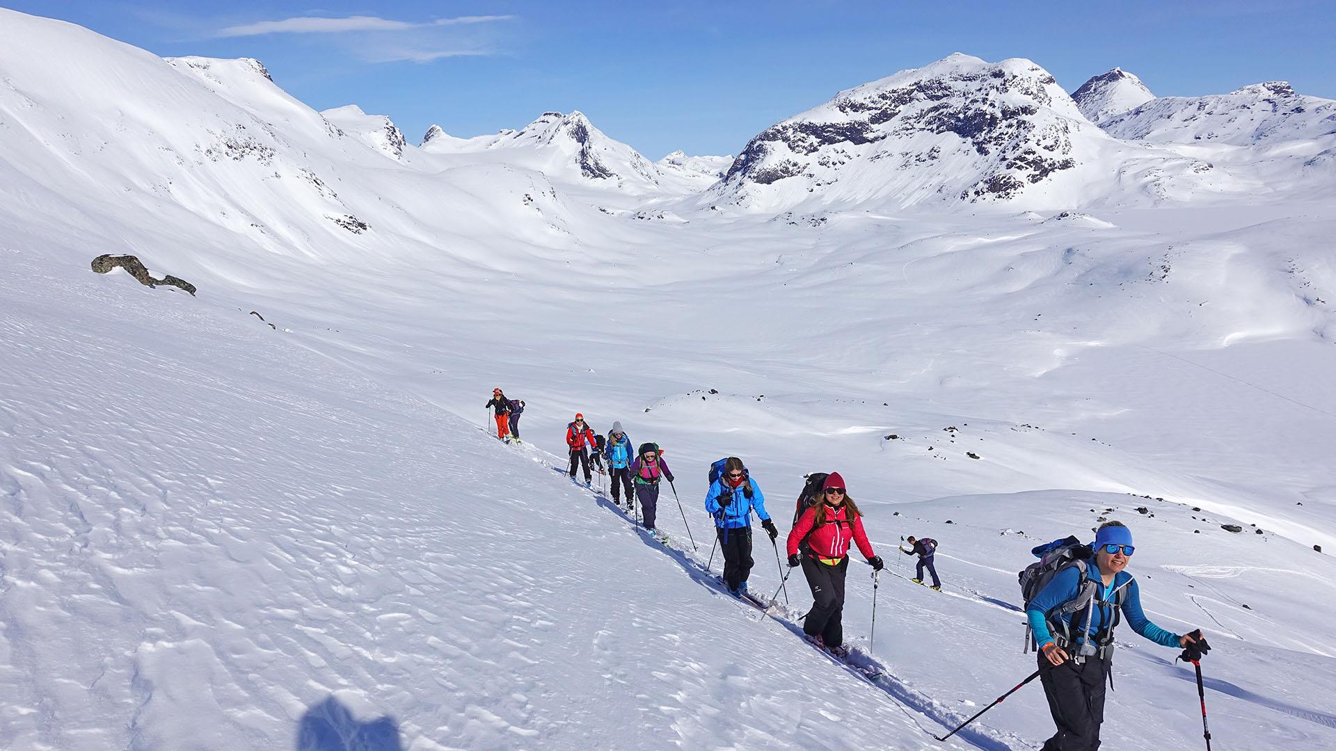 Eine Gruppe von Bergskiläufern in einer Reihe in einer Bergflanke mit hohen Bergen im Hintergrund.