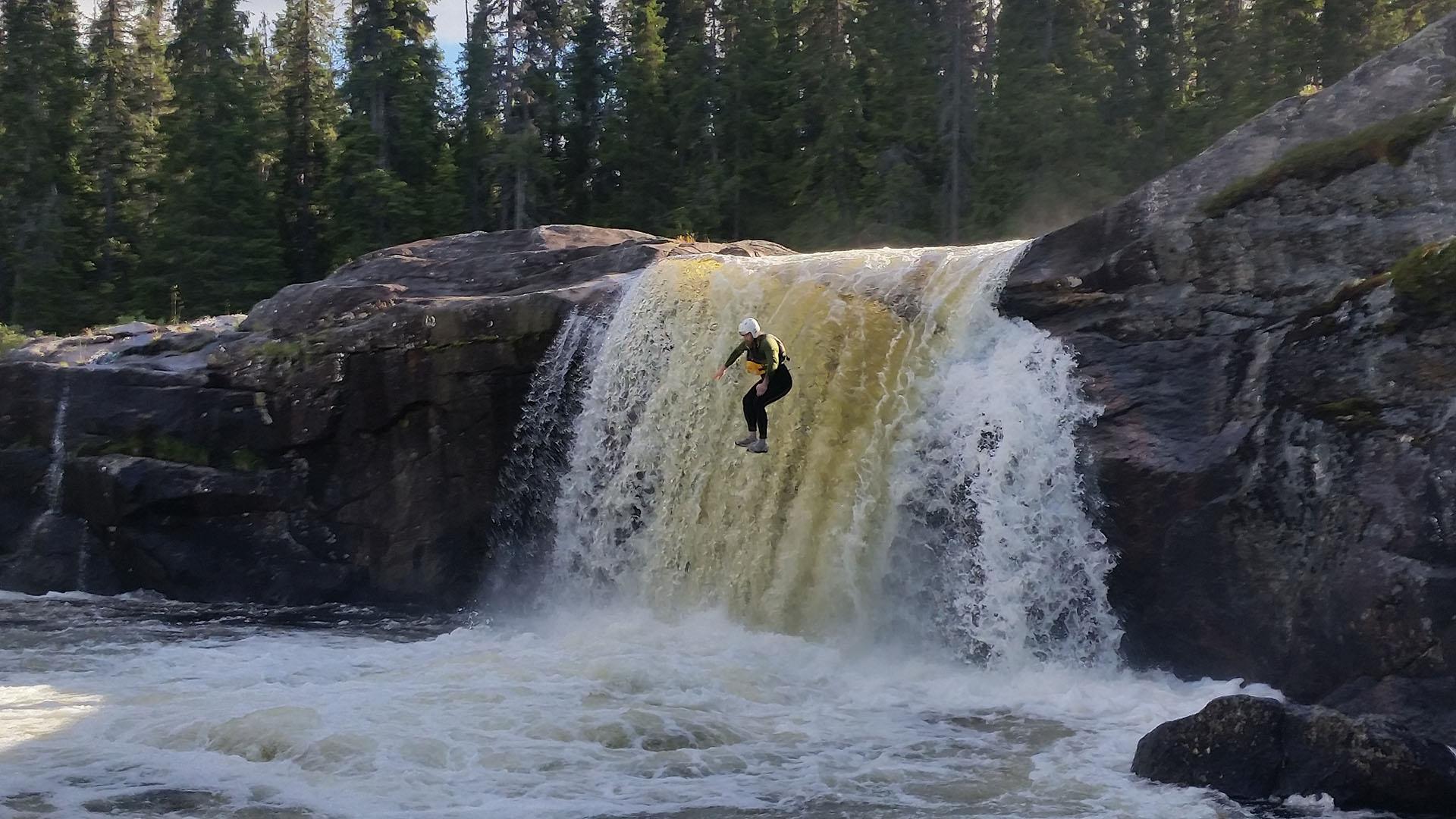 A man in wetsuit and helmet jumps down a waterfall in a river.