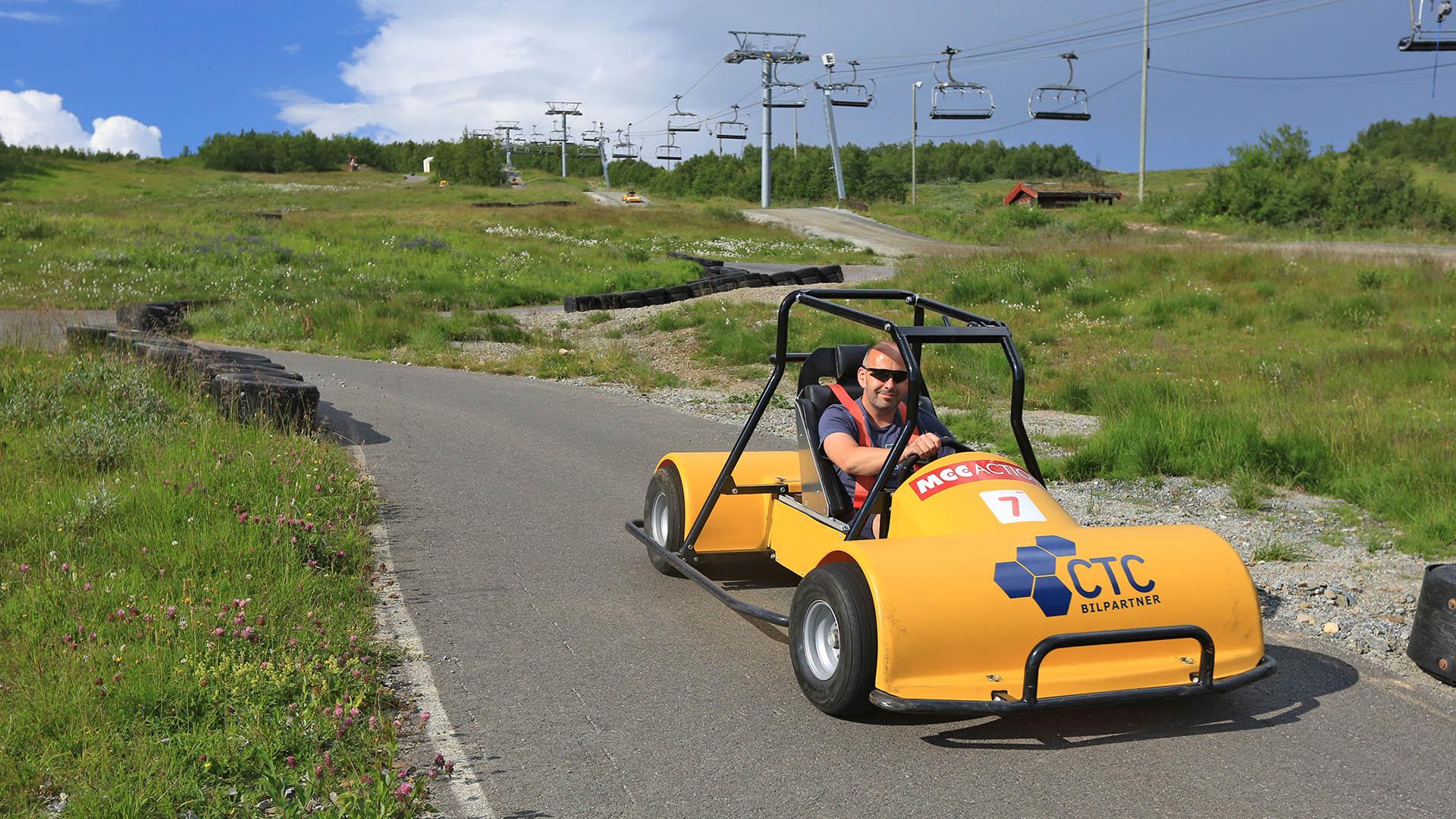 A man steers a yellow go-cart in a paved downhill-track. A summer ski lift can be seen in the background.