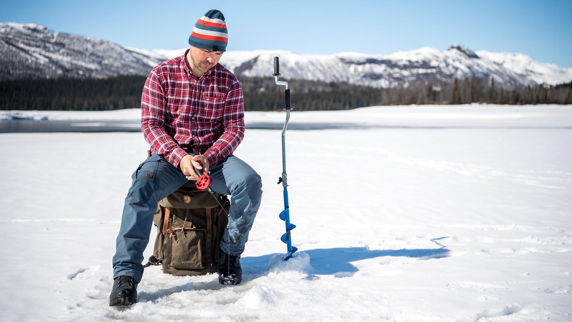 Ice fisherman in red-checked flannel sits on a ice-covered lake with mountains in the background.
