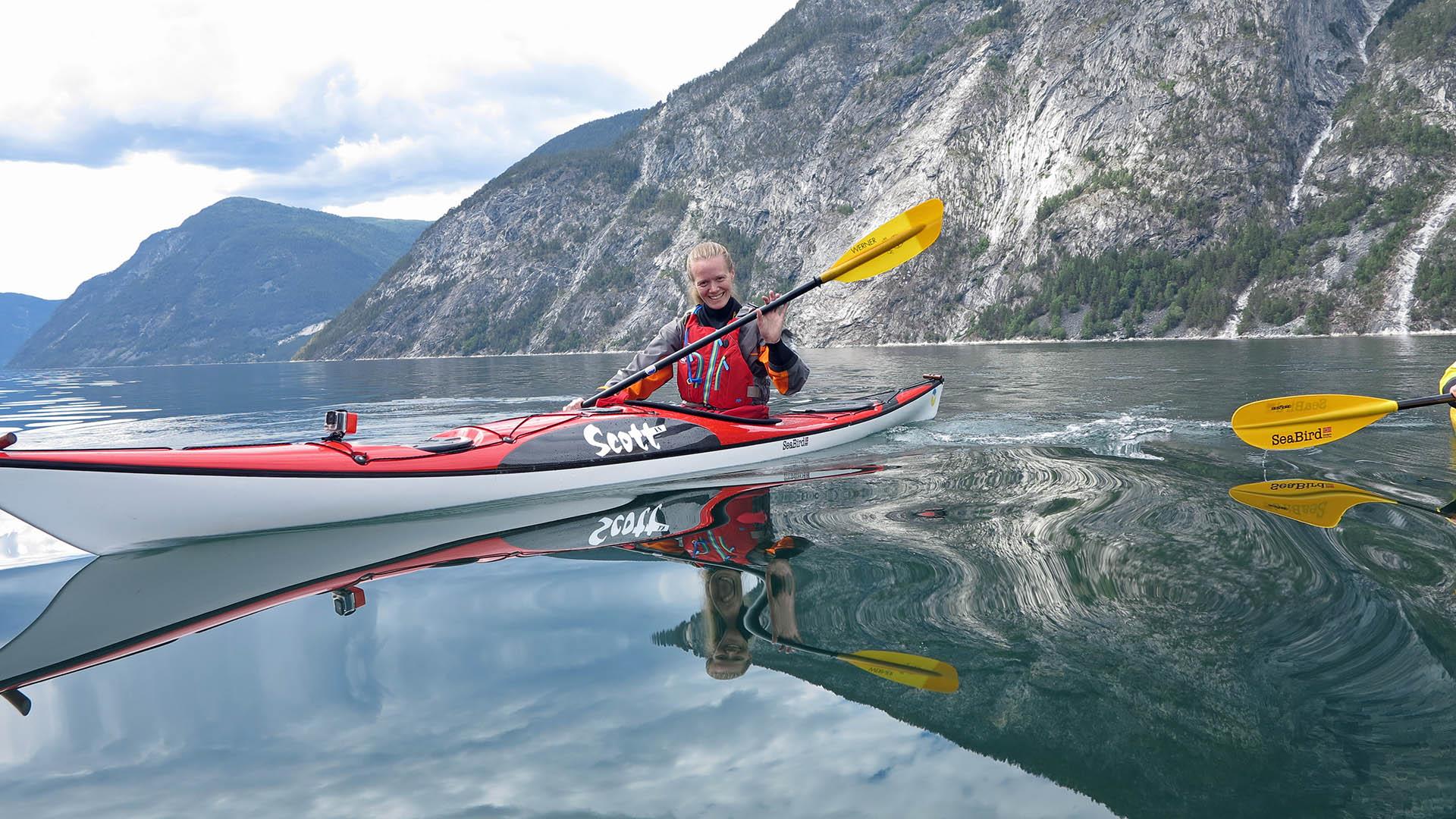 A young lady with red life evst in a red-grey kayak with yellow paddle blades on a mirror-calm fjord.