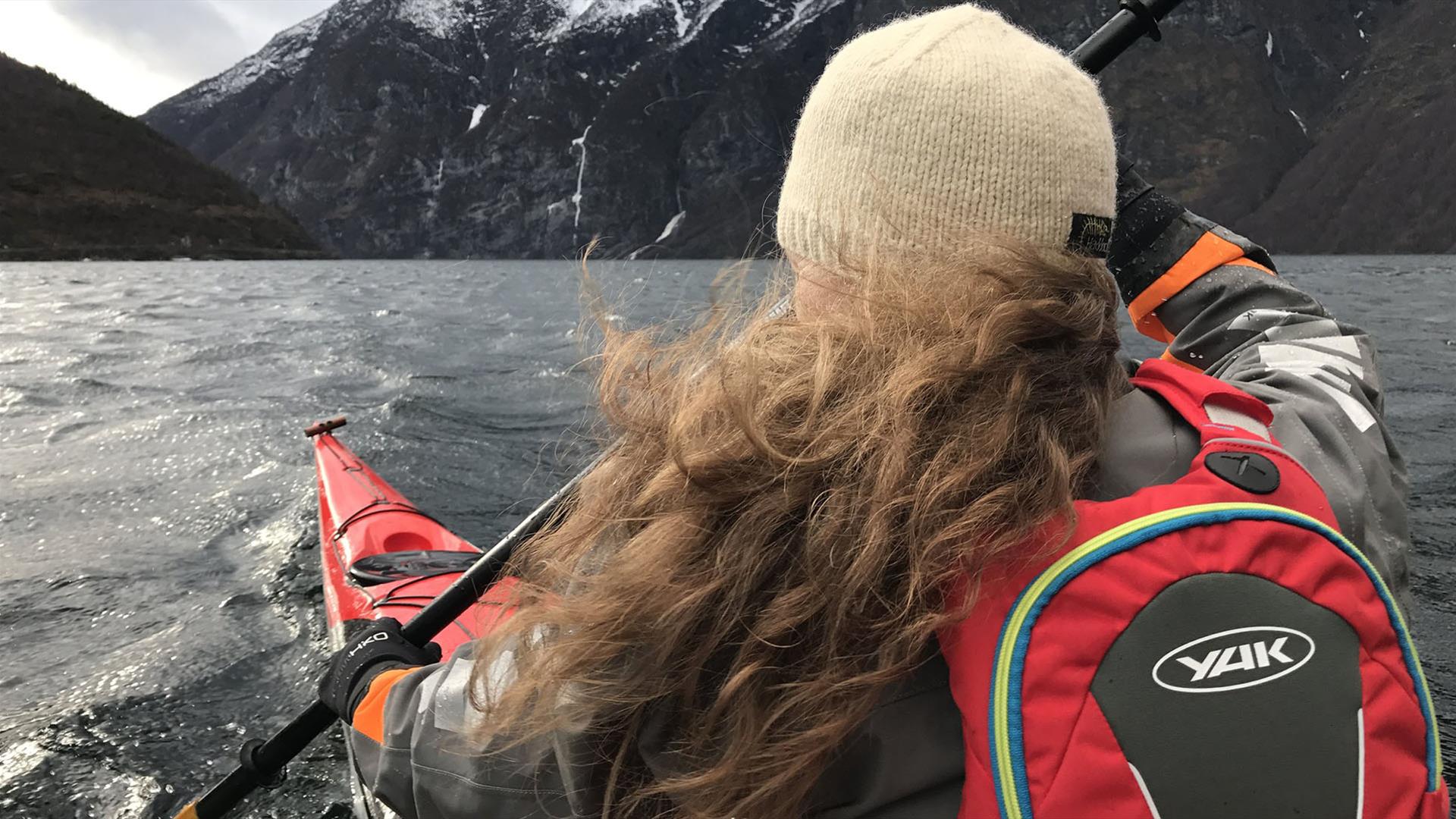 A lady with long curly hair and a woolen hat in a red kayak on a fjord in windy and cold weather.