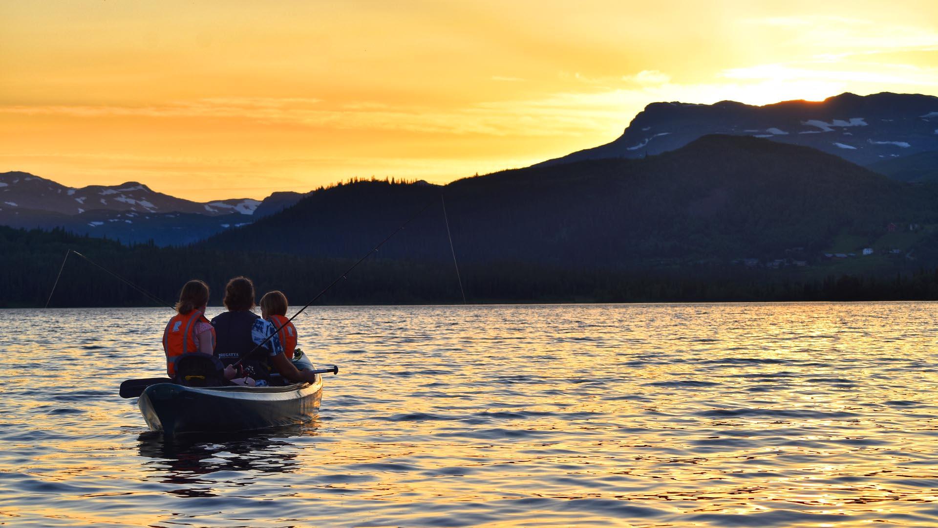 A canoe with three persons that fish with rod on a lake with mountains in teh background. The setting sun paints the sky and the water in yellow and orange.
