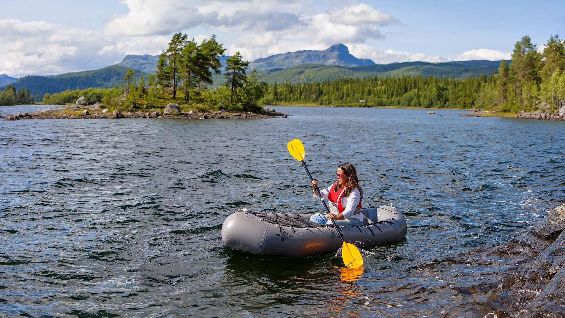 En person i en packraft med gul paddelåre på et fjellvann. En liten øy med furutrær og et fjell ses i bakgrunnen.