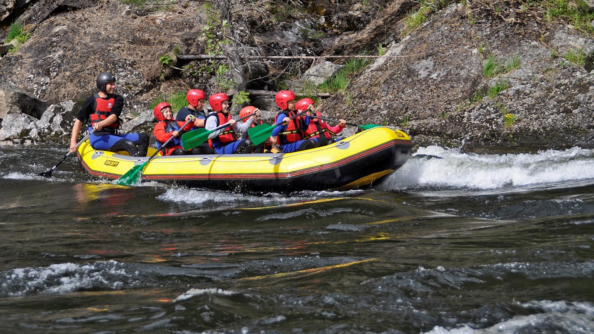 Ein Raftingboot mit Führer und Kindern auf einem Fluss mit ruhigen Stromschnellen und Felsenufer.