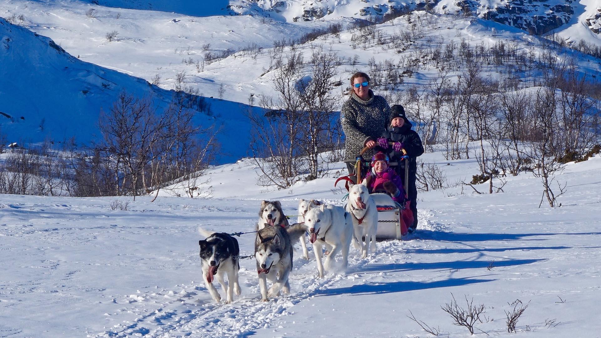 Ein Hundeschlittengespann mit zwei Kindern, die als Passagiere den Schlittenführer begleiten, kommt in einer verschneiten Landschaft auf den Fotografen zugelaufen.