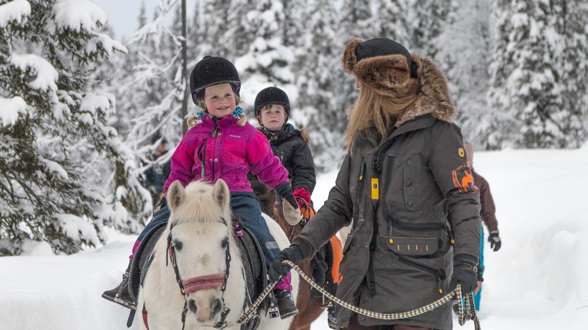 A woman with a fur hat leads two ponies with children riding