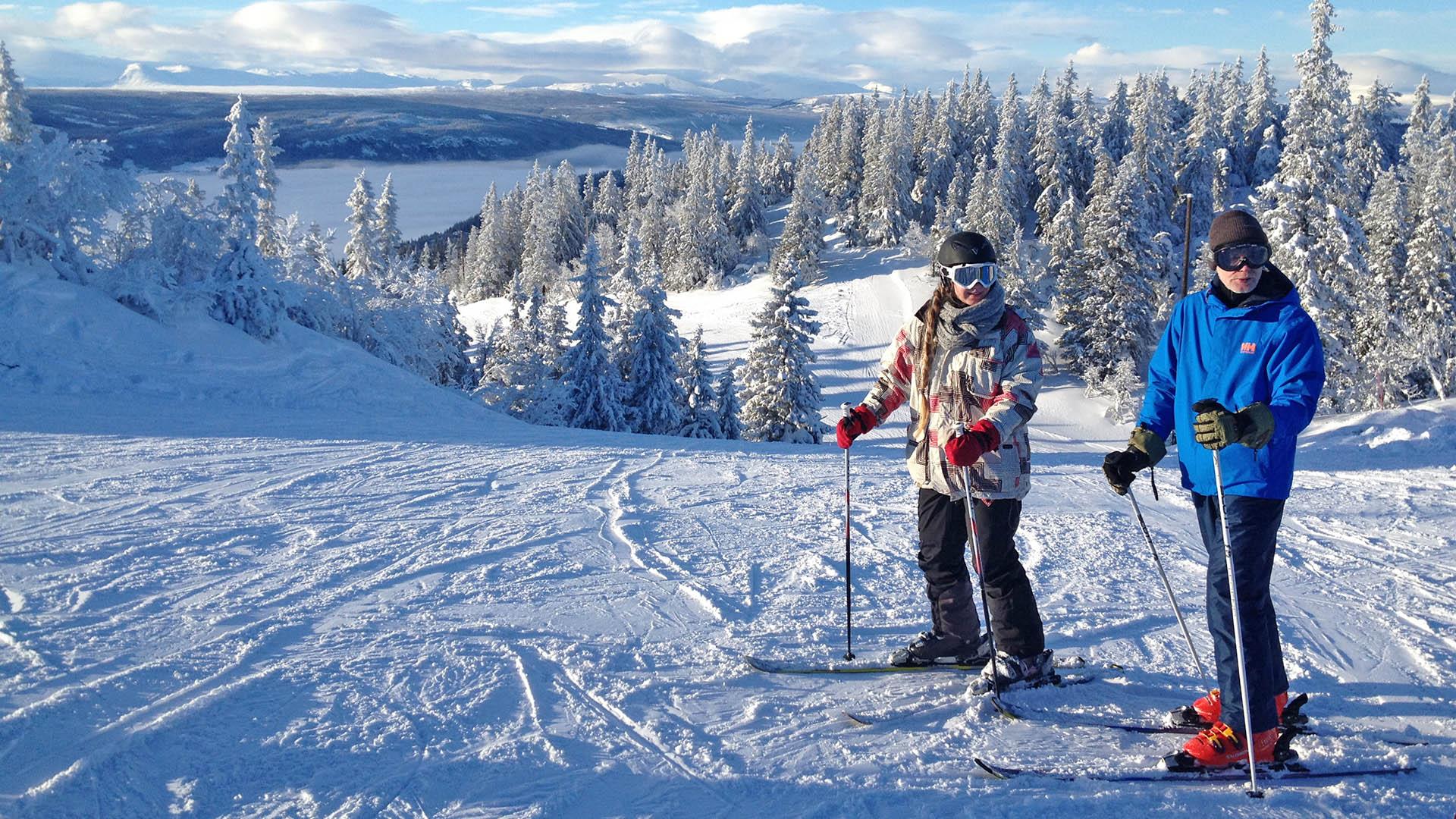 Zwei Skifahrer auf der Piste schauen zum Fotografen. Schnee liegt auf den Fichten im Hintergrund.
