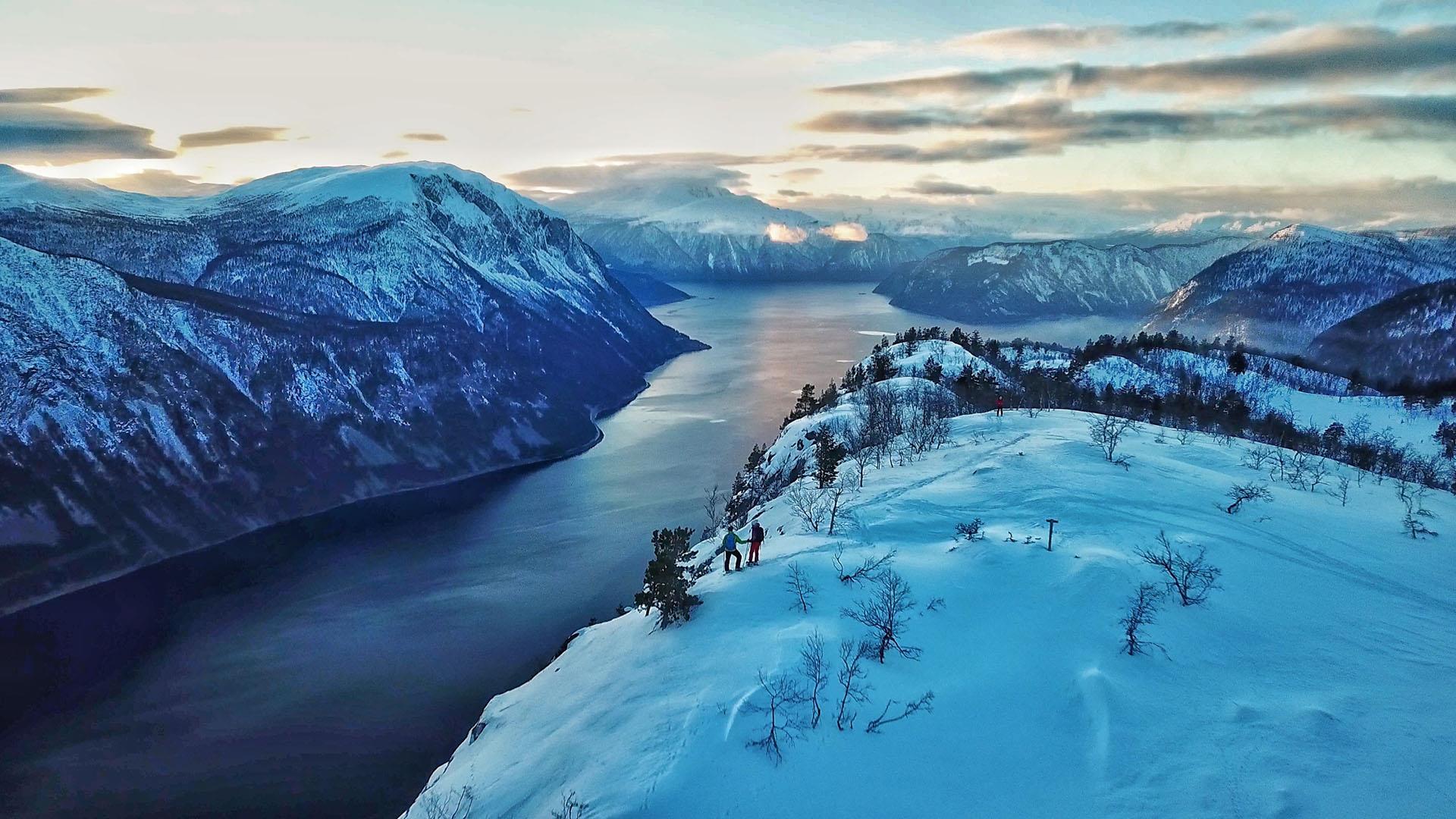 Fjellvandrere på truger i et nedsnødd fjord- og fjellandskap i magisk vinterlys.