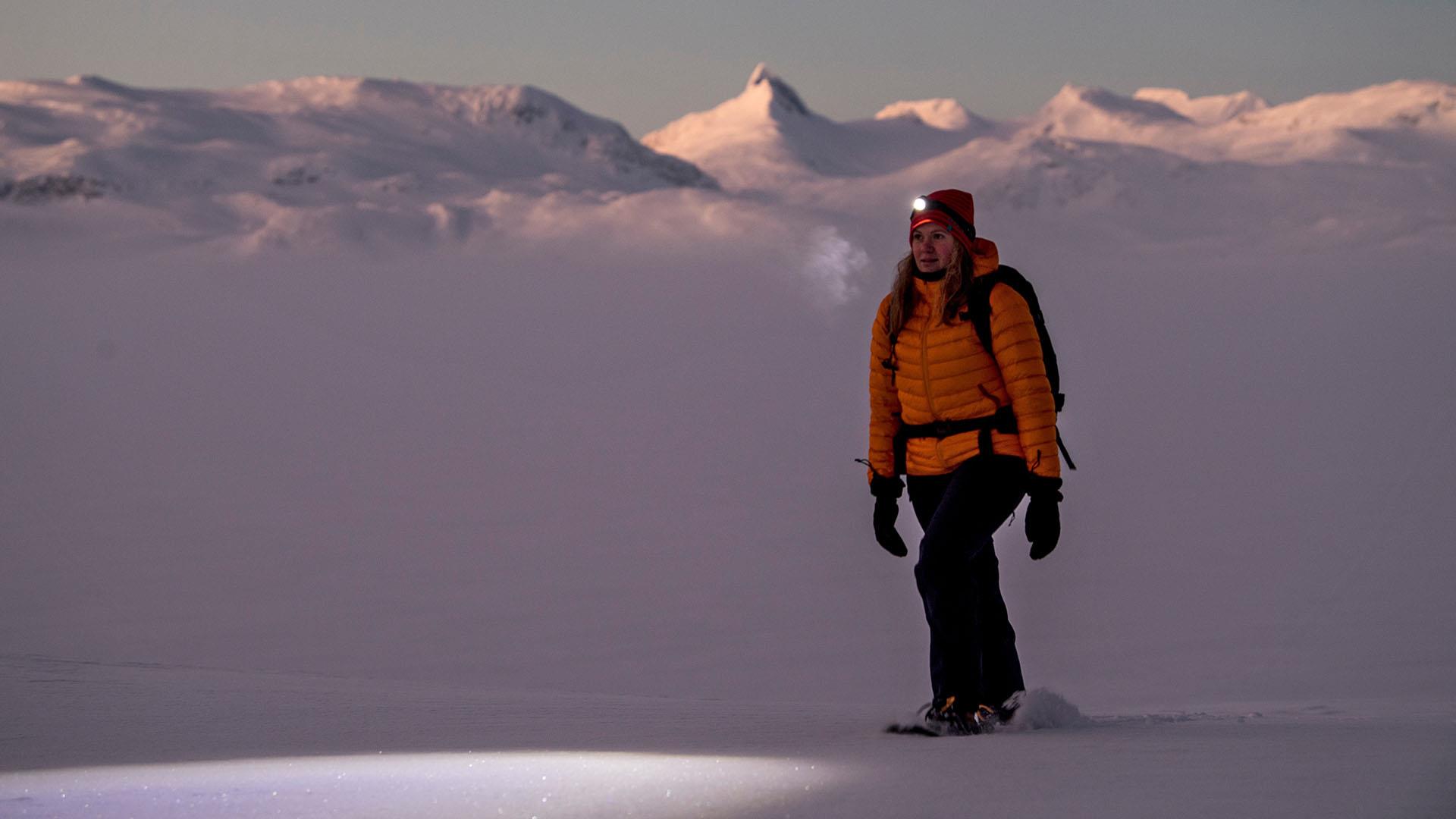 A woman in winter clothes and a head lamp walks on snowshoes in the twilight in the mountains. Some high, pointed peaks rise in the background.
