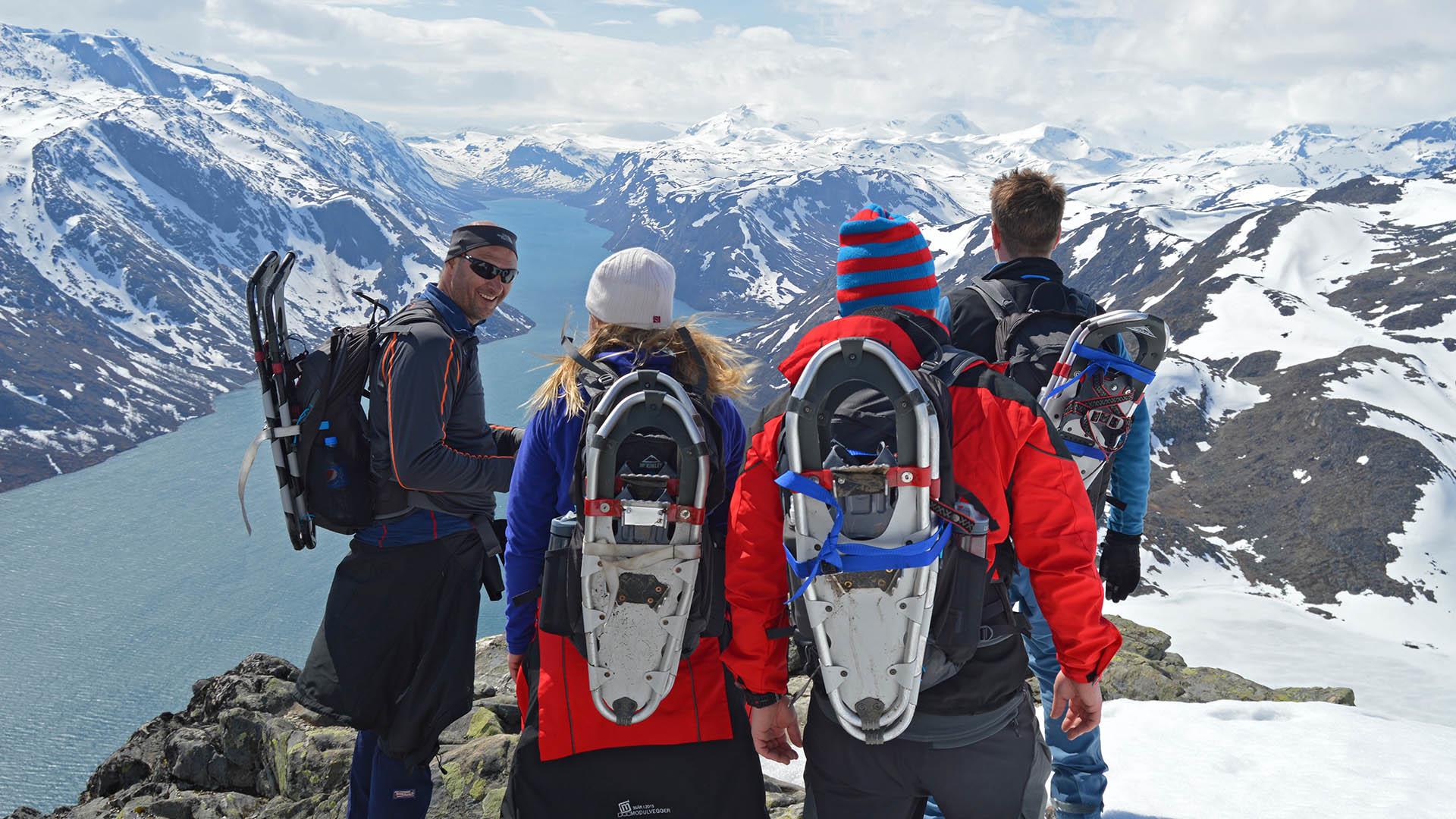 A group of hikers with snowshoes strapped to their backpacks in the high mountains with lakes and summits early in the summer when there is still much snow left.