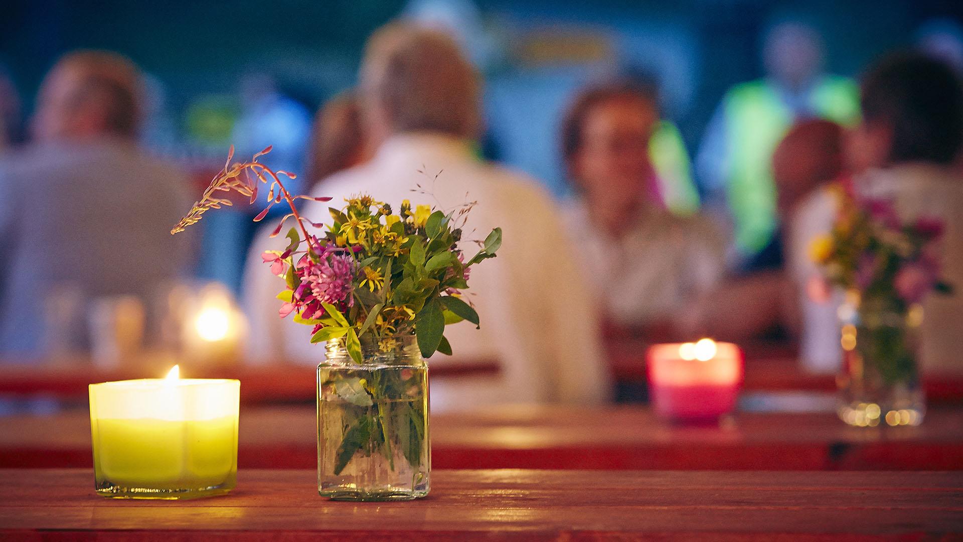 Wiesenblumen in einem Glas und Teelichter auf einem Tisch im Vordergrund an einem lauen Sommerabend, mit heiter gestimmten Leuten unscharf im Hintergrund.