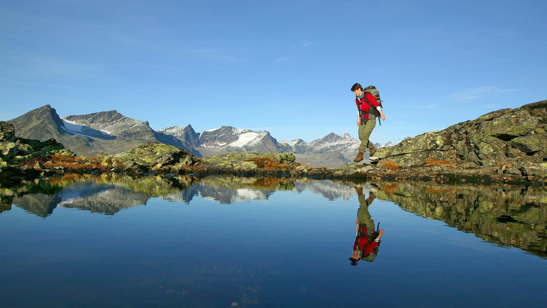 A woman and her reflection as she walks over rocks behind a small pond with a completely calm water surface. In the background high summits rise.