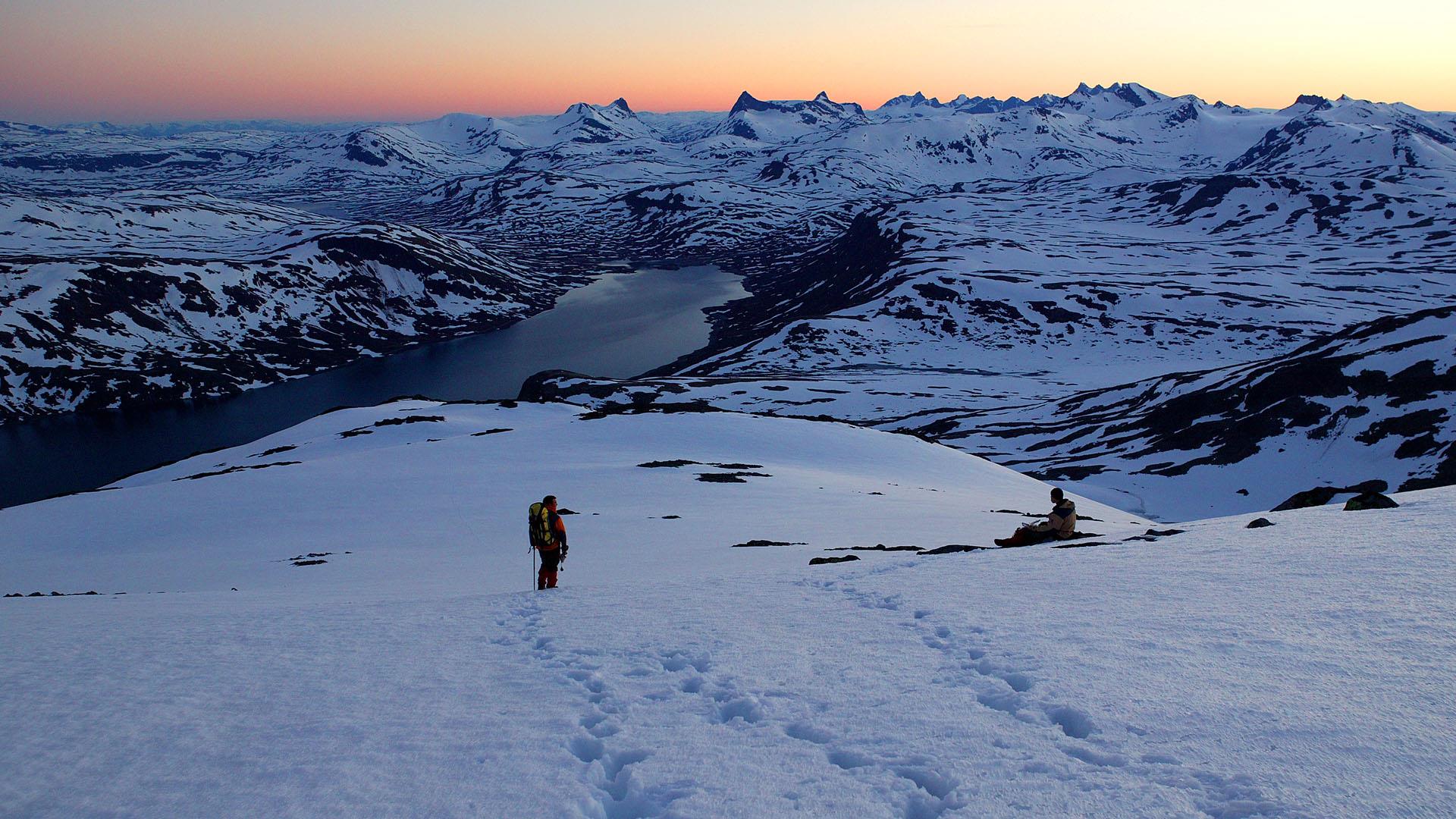 Zwei kleine Personen in einer mächtigen Berglandschaft mit einem See und vielen spitzen, hohen Bergen vor Sonnenaufgang. Der Horizont ist vom Morgenrot gefärbt, und es ist Frühsommer, und es liegt noch viel Schnee.