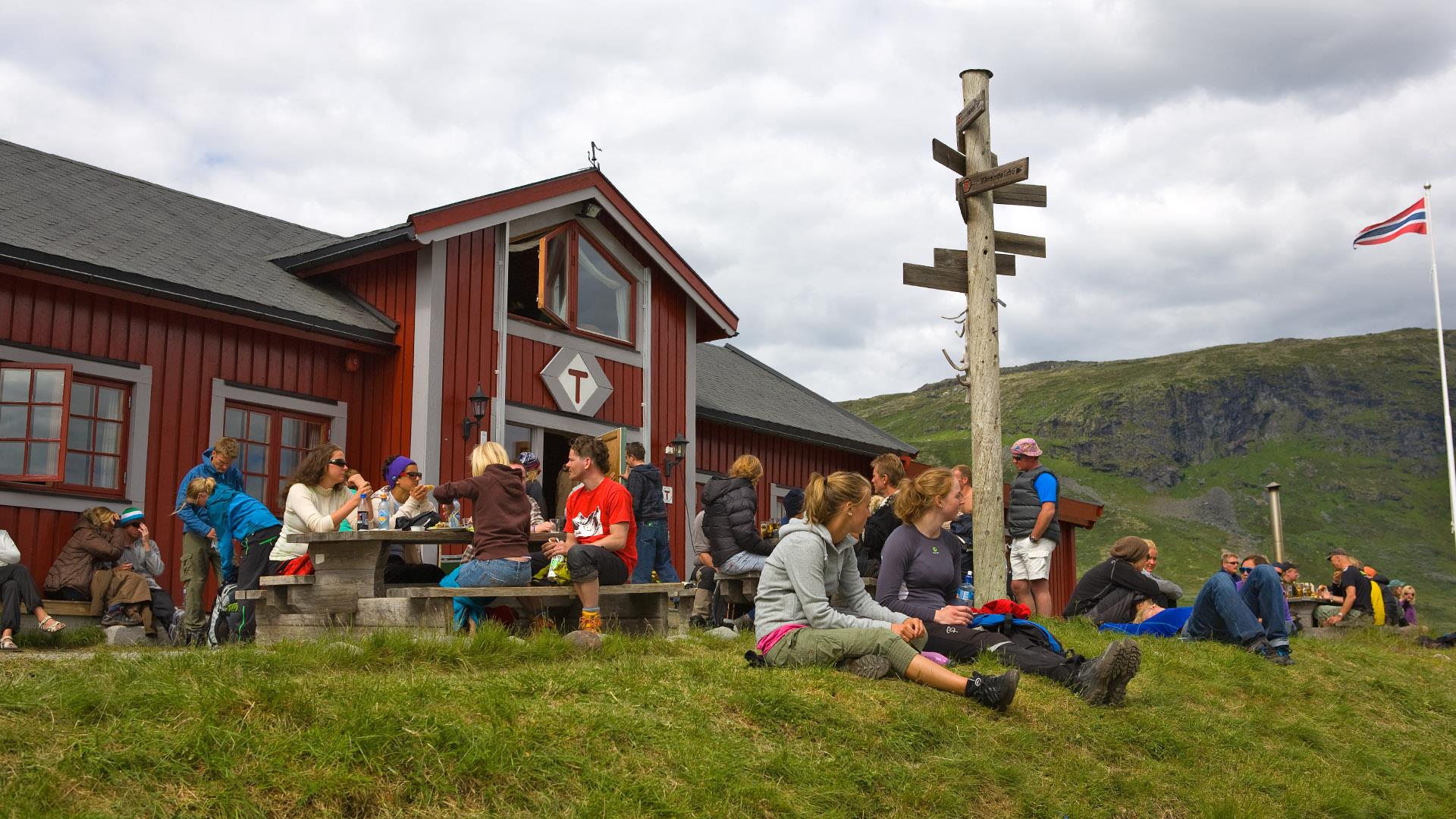 Mountain hikers relax outside the tourist cabin Fondsbu, a red wooden buiding with the logo of the Norwegian Mountain Association (DNT) over the entrance