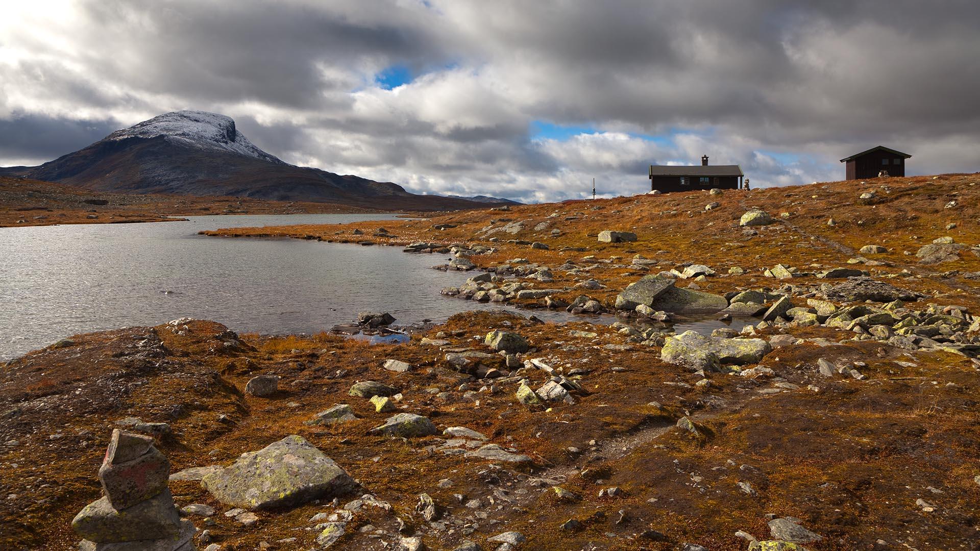 Two cabins by a lake in open highland country with a mountain in the background. It is autumn, the ground is orange-brown-coloured and snow has been falling on the mountain top.