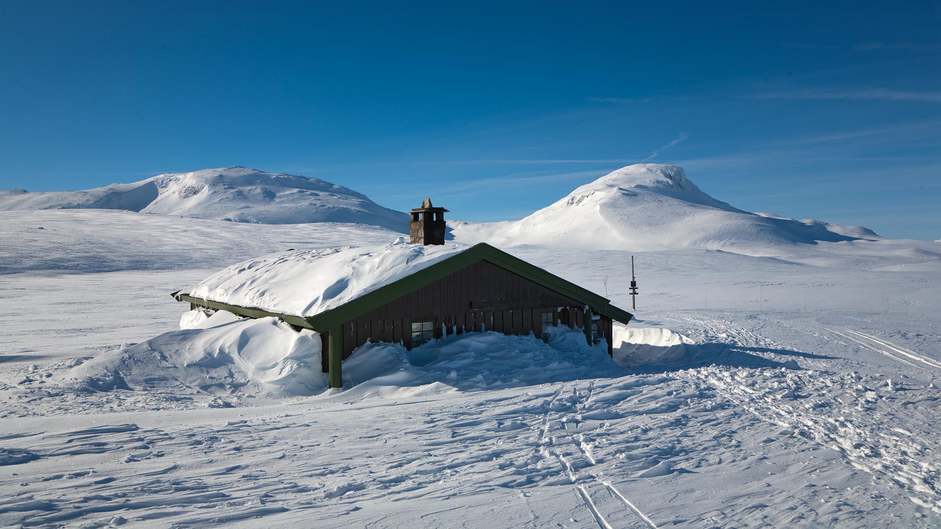 A lone cabin with snow up under the roof and two mountain tops in the background. Ski tracks lead to the entrance which has been dug out.