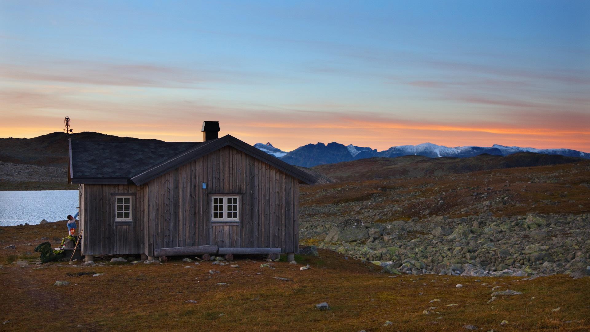 Eine Hütte im Fjell an einem See. Der Sonnenuntergang färbt den Himmel über einer Reihe spitzer Berggipfel am Horizont orange.