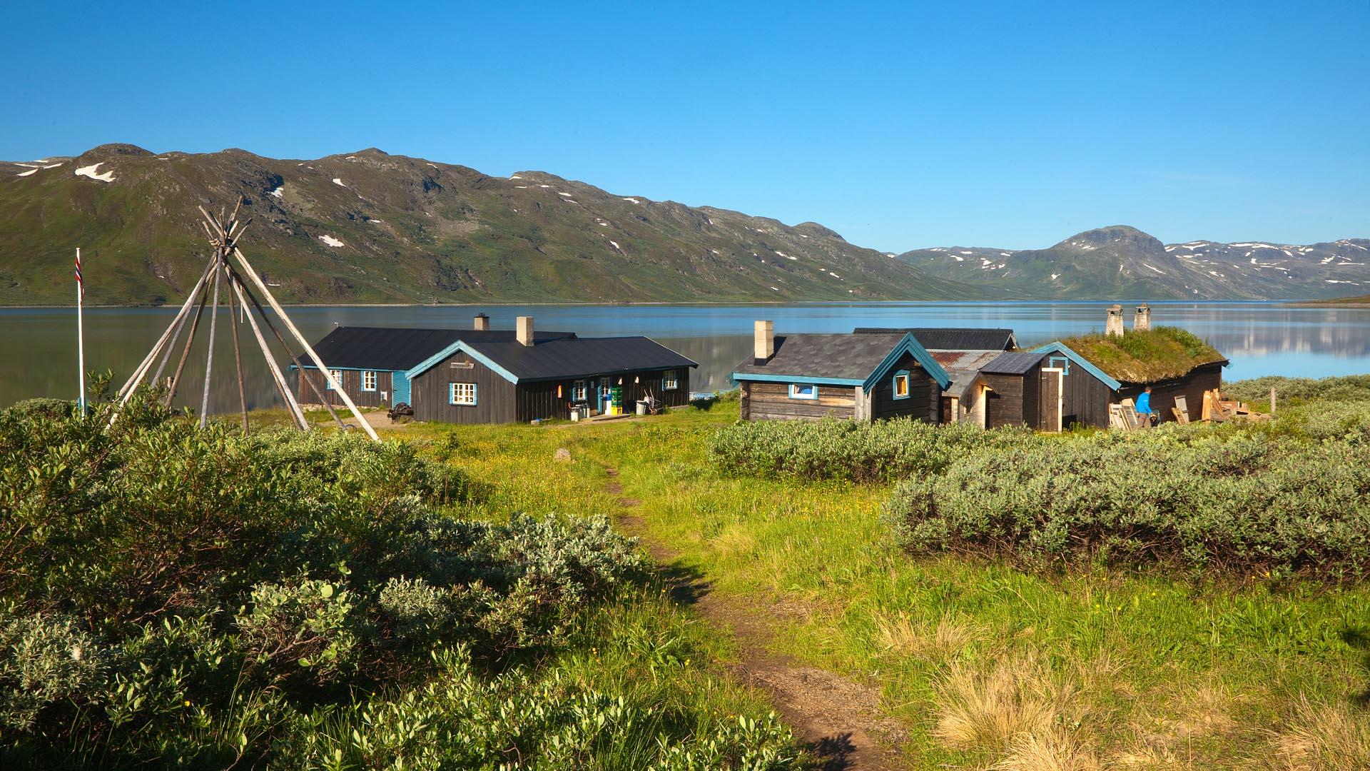 A grassy trail leads through juniper bushes to a compund of cabins on the shore of a large mountain lake.