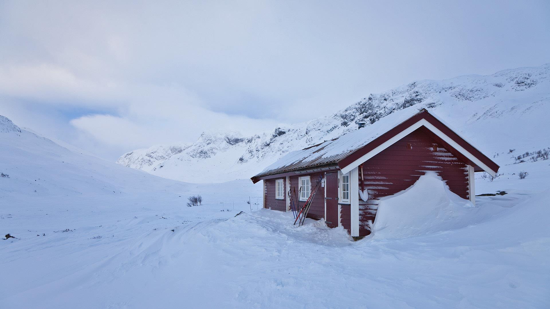 En mørkrød hytte i en fjelldal om vinteren. Flatt lys og grå himmel.