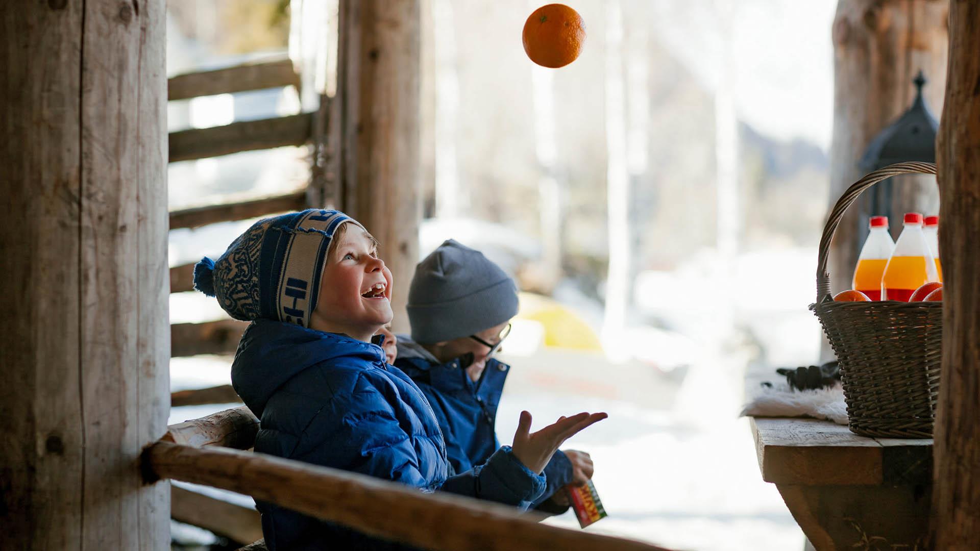 Jungs in Winterkleidung jonglieren mit einer Apfelsine. Auf dem Tisch ein Korb mit Brauseflaschen.