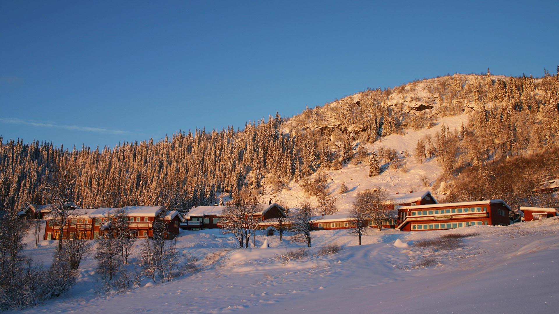 Lange, niedrige rotgemalte Holzgebäude am Fuße einer Anhöhe in offenem Gelände, umgeben von Fichtenwald in weichem Wintersonnenlicht. Es liegt Schnee, auch auf den Bäumen.