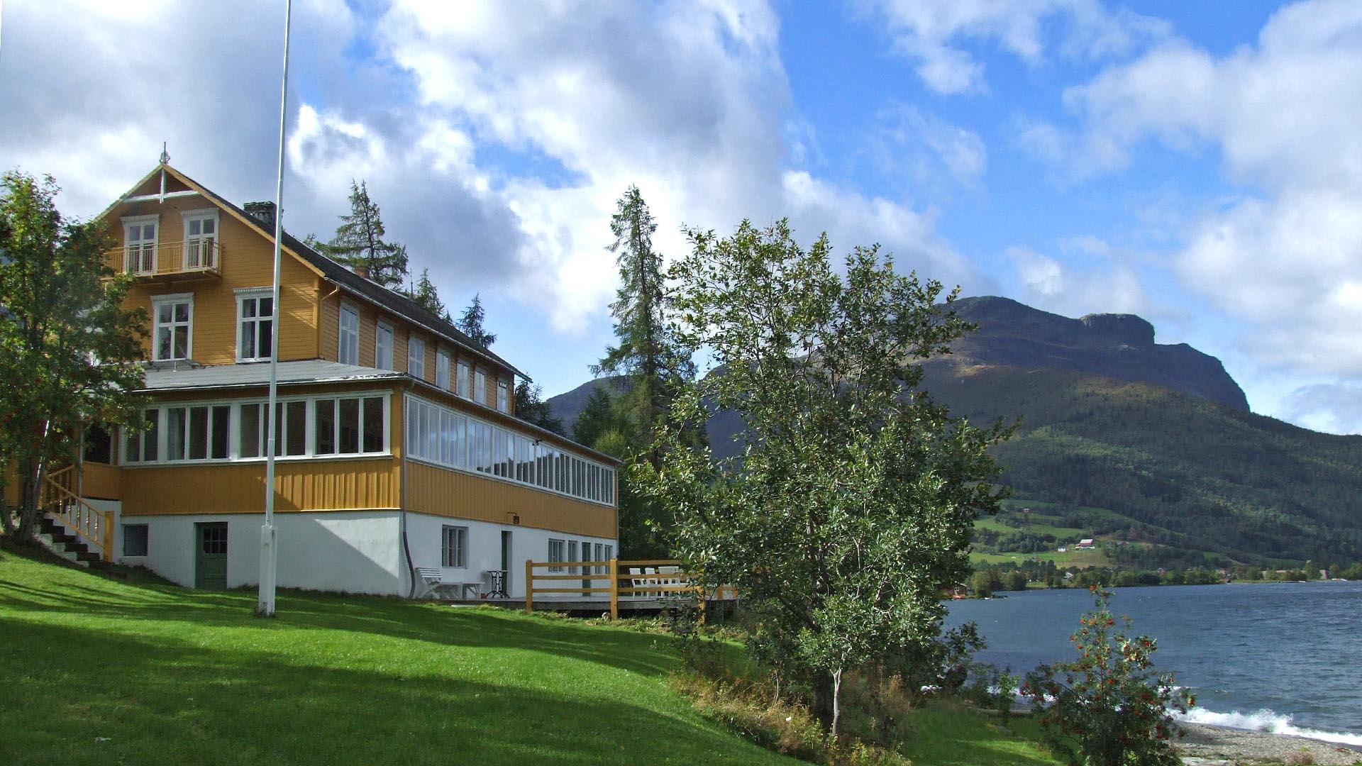 A charming yellow-painted small wooden hotel on a lakefront with a lawn in the fore- and a mountain in the background