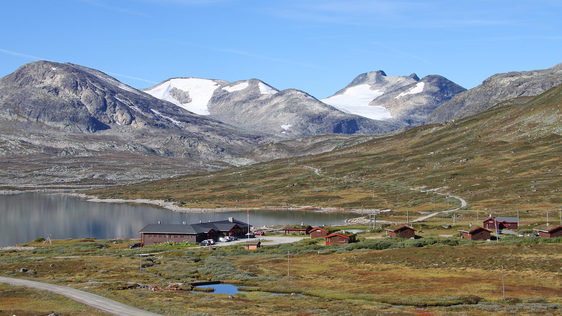 Eine Lodge und Hütten liegen am Ufer eines Bergsees in offener Fjellandschaft mit hohen, schneebedeckten Bergen im Hintergrund