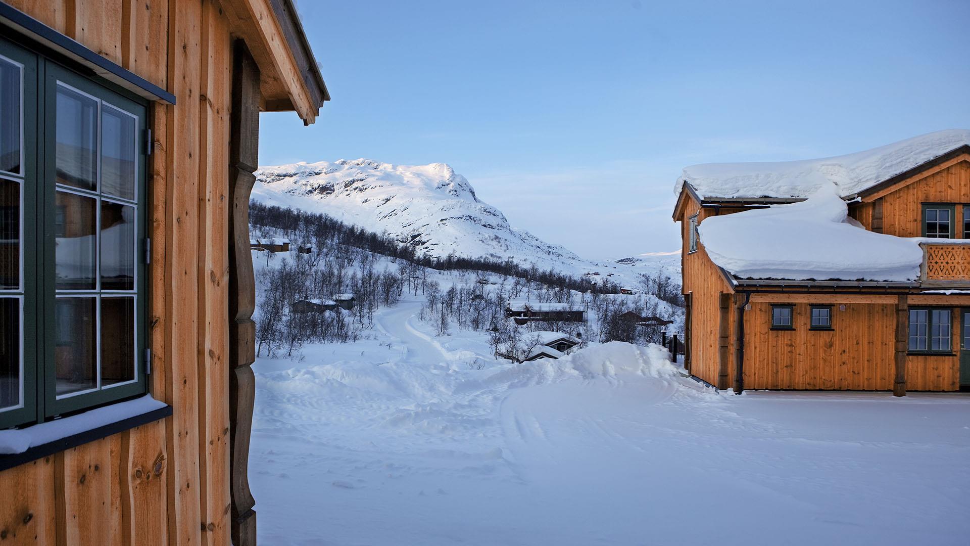 Utsikt mellom to hyttevegger mot et fjell der kveldssola så vidt fortsatt skinner på toppen. Vinter og snø.