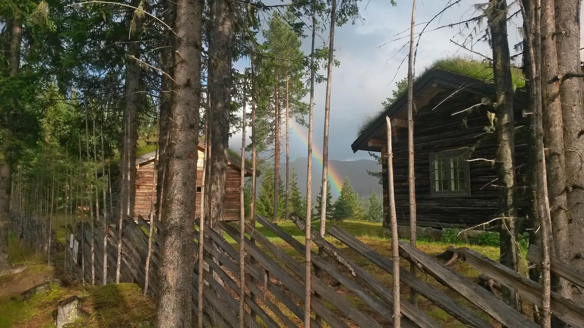 Round pole fence in front of old log cabin with grass roof and a rainbow