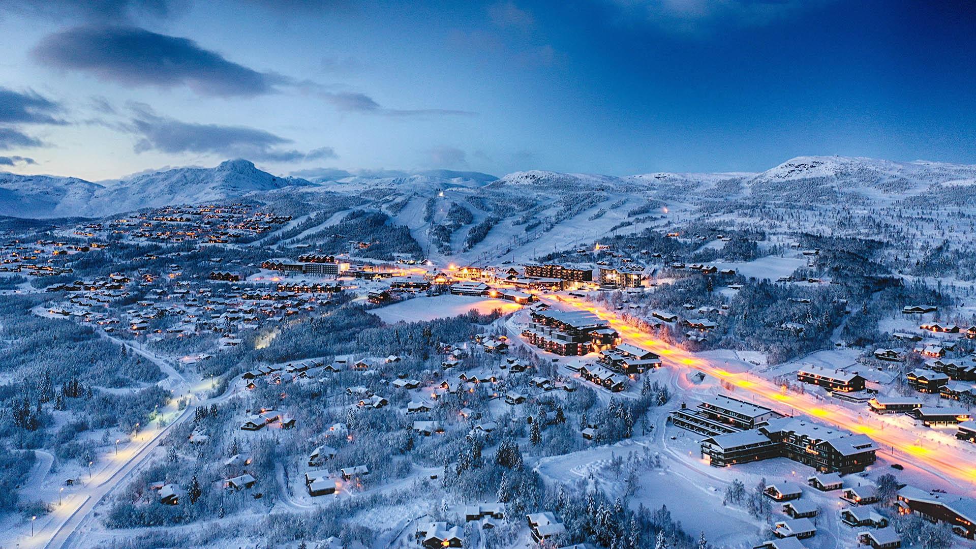 Dronebilde utover en snødekt fjellandsby i blåtimen en vinterkveld med en vei som er opplyst av gatelys, en alpinbakke og fjell i den fjerne horisonten
