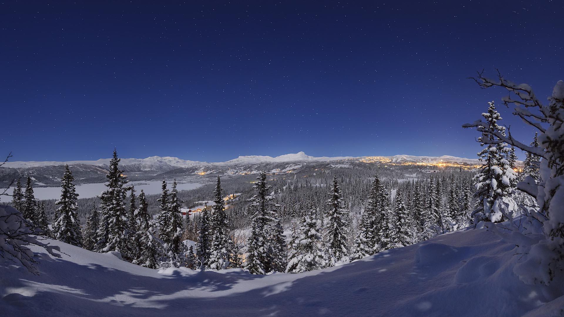 Weitwinkelaufnahme bei Nacht von einer Anhöhe über verschneiten Fichtenwald zu einem Bergdorf, dessen Lichter am Horizont leuchten.