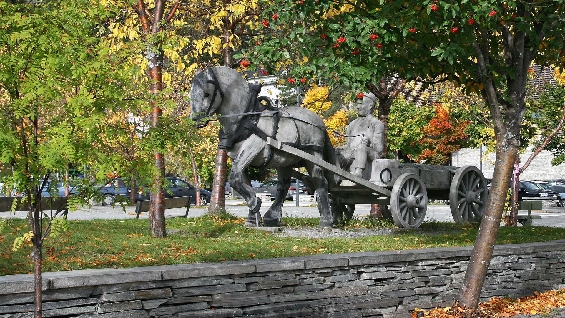 En plenstripe på toppen av en lav skifermur, me en steinskulptur av en hest med kusk og vogn som er lastet med skifer. Trær i høstdrakt står omkring.