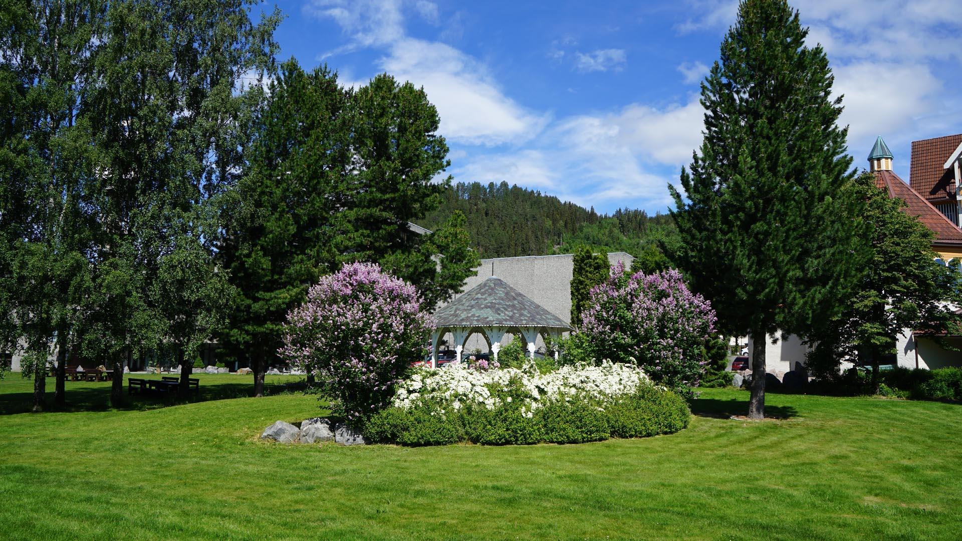 I en park med plen og blomsterbed og blomstrende syrinbusker rundt en hagepaviljong. I bakgrunnen står noen høye trær.
