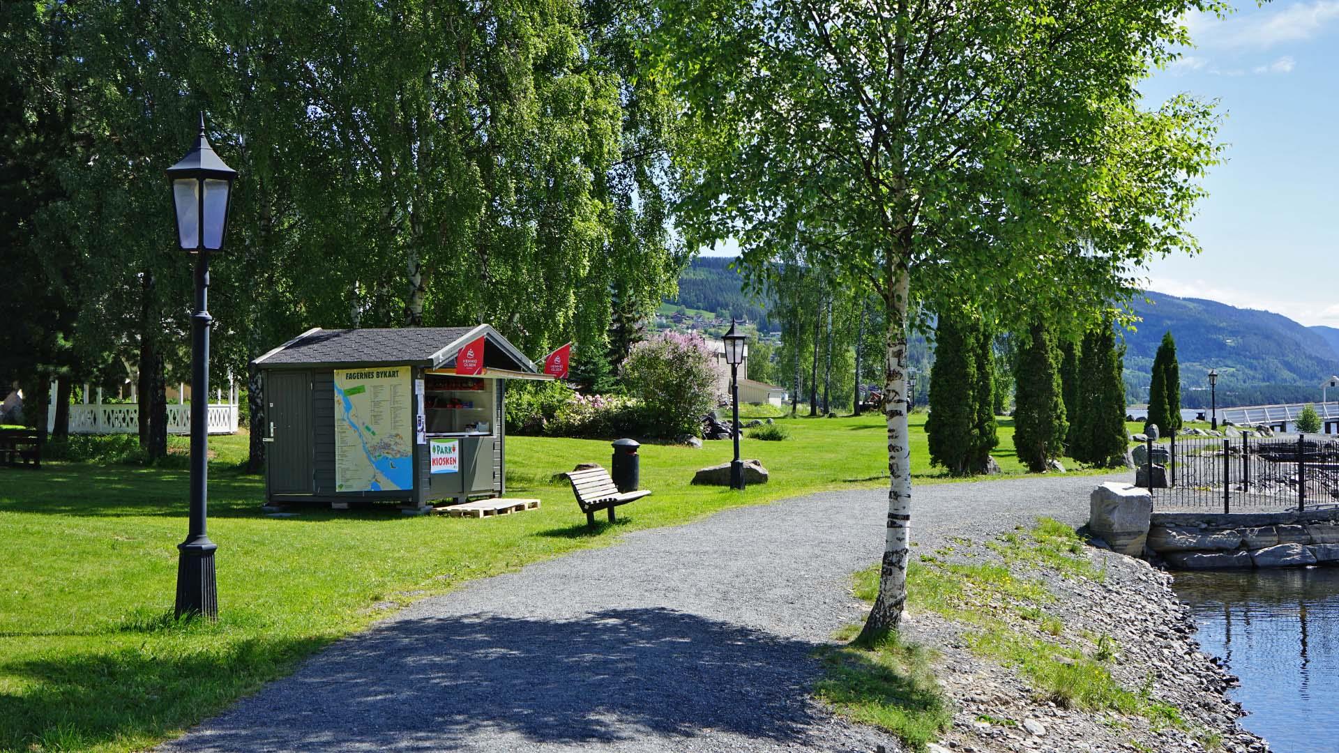 Strandpromenade langs Strandefjorden i parken på Fagernes om sommeren med iskiosk, gatelykt, benk, grønn plen og bjørketrær