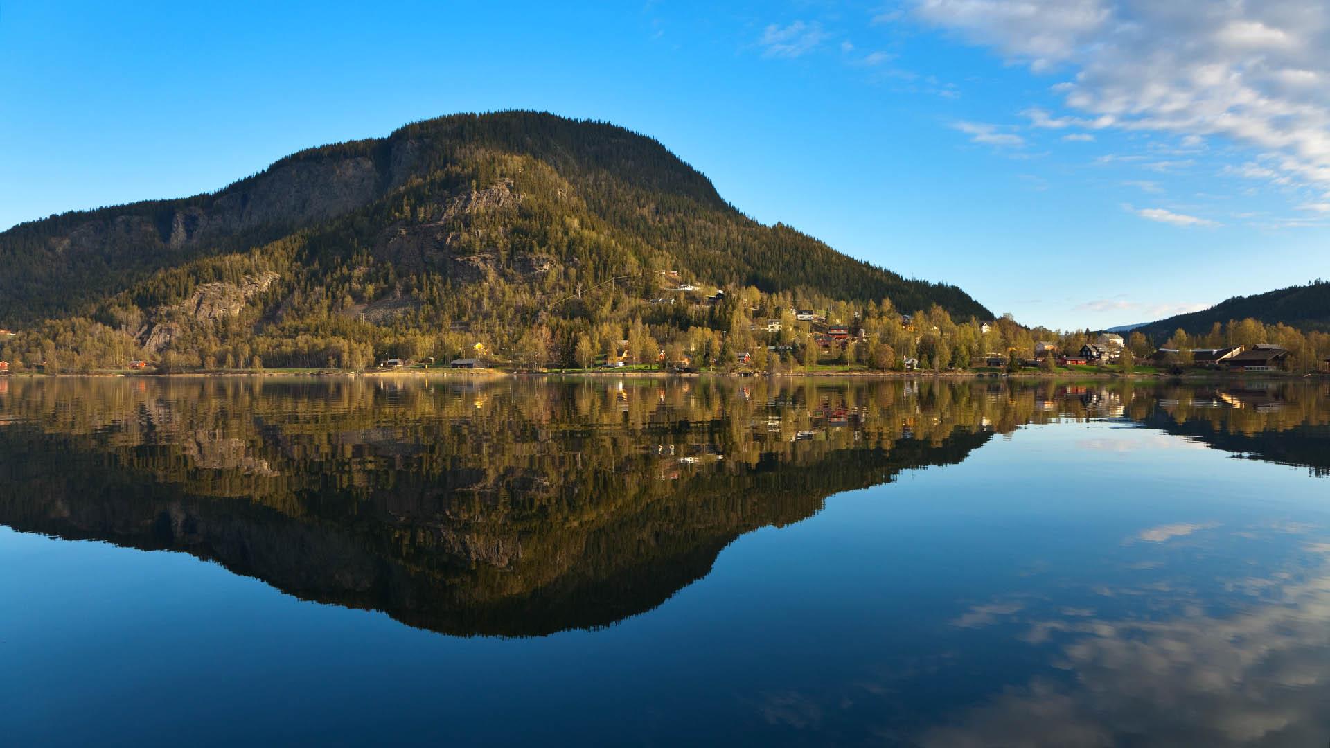 The small town of Fagernes seen from the lake, with a forested hill beyond the town that is mirrored on the blank surface of the water. Blue sky.
