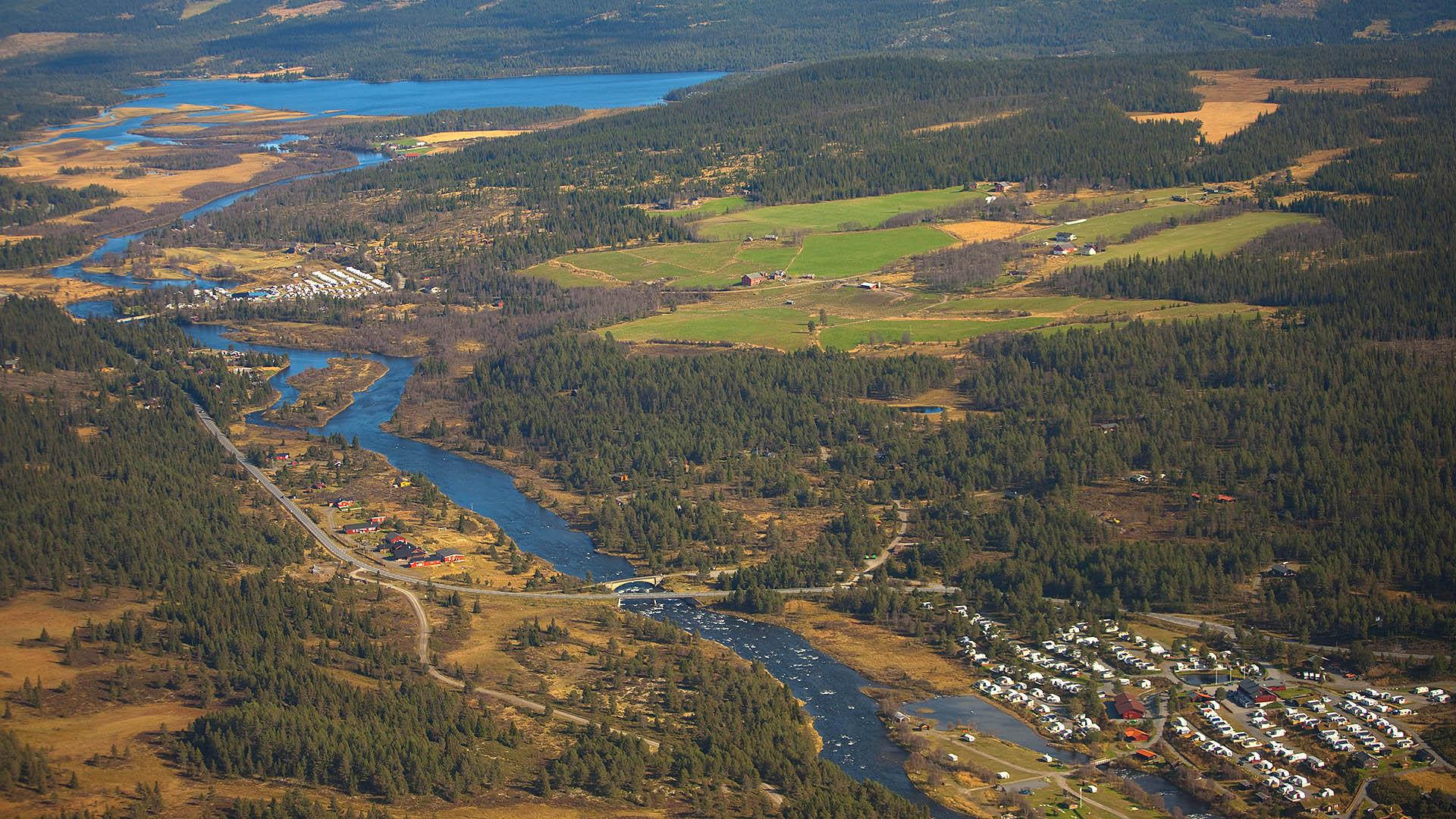 Luftaufnahme eines breiuten Talbodens mit einem Fluss, der an einem Campingplatz, ein paar Waldstücken und ein paar grünen Weiden vorbeifließt. Im Hintergrund ein See. Sommer.