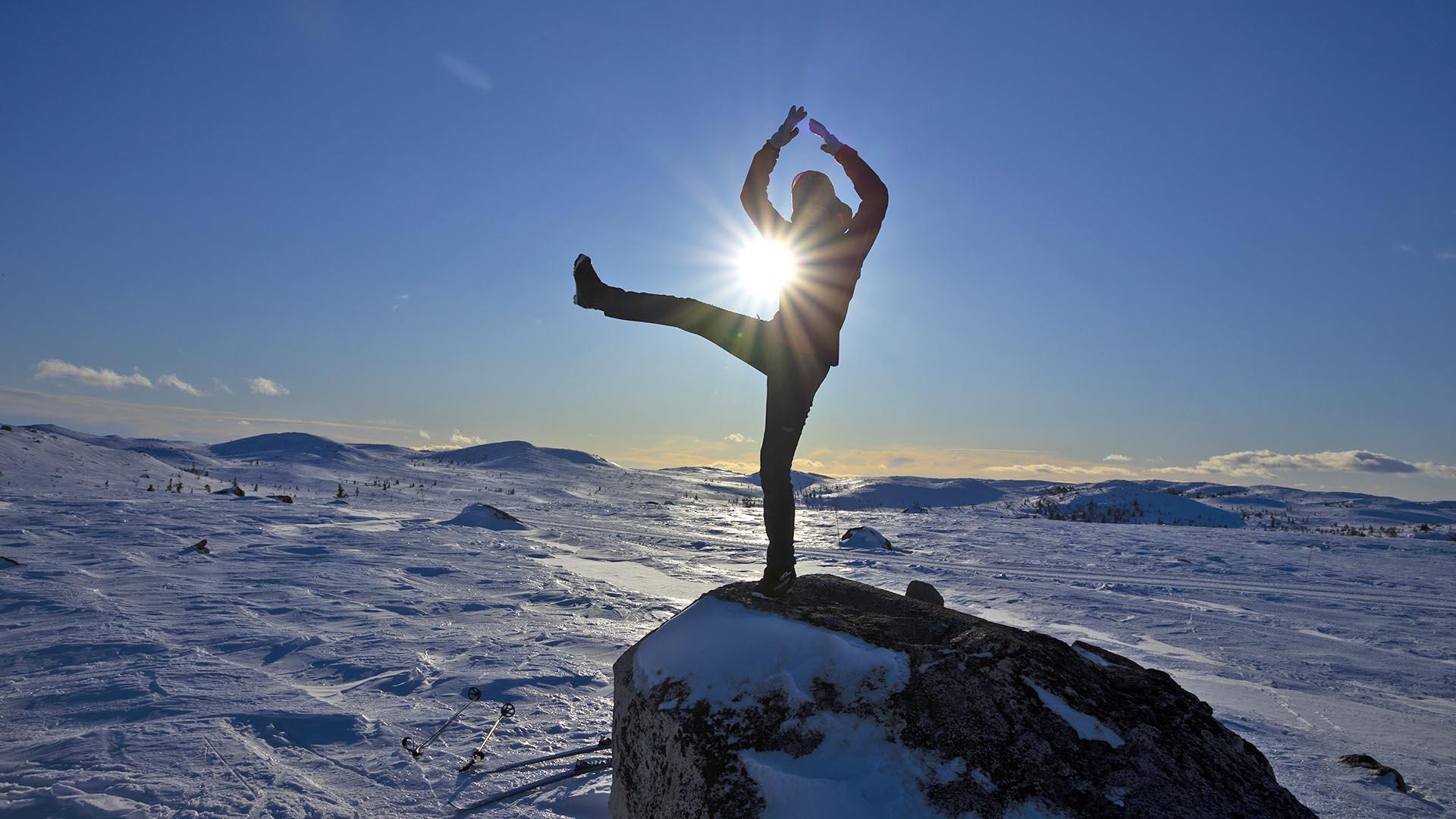 En jente på en steinblokk i motsol på vinterfjellet. Hun poserer slik at sola danner en mangestrålet stjerne mellom et hevet bein og en arm.
