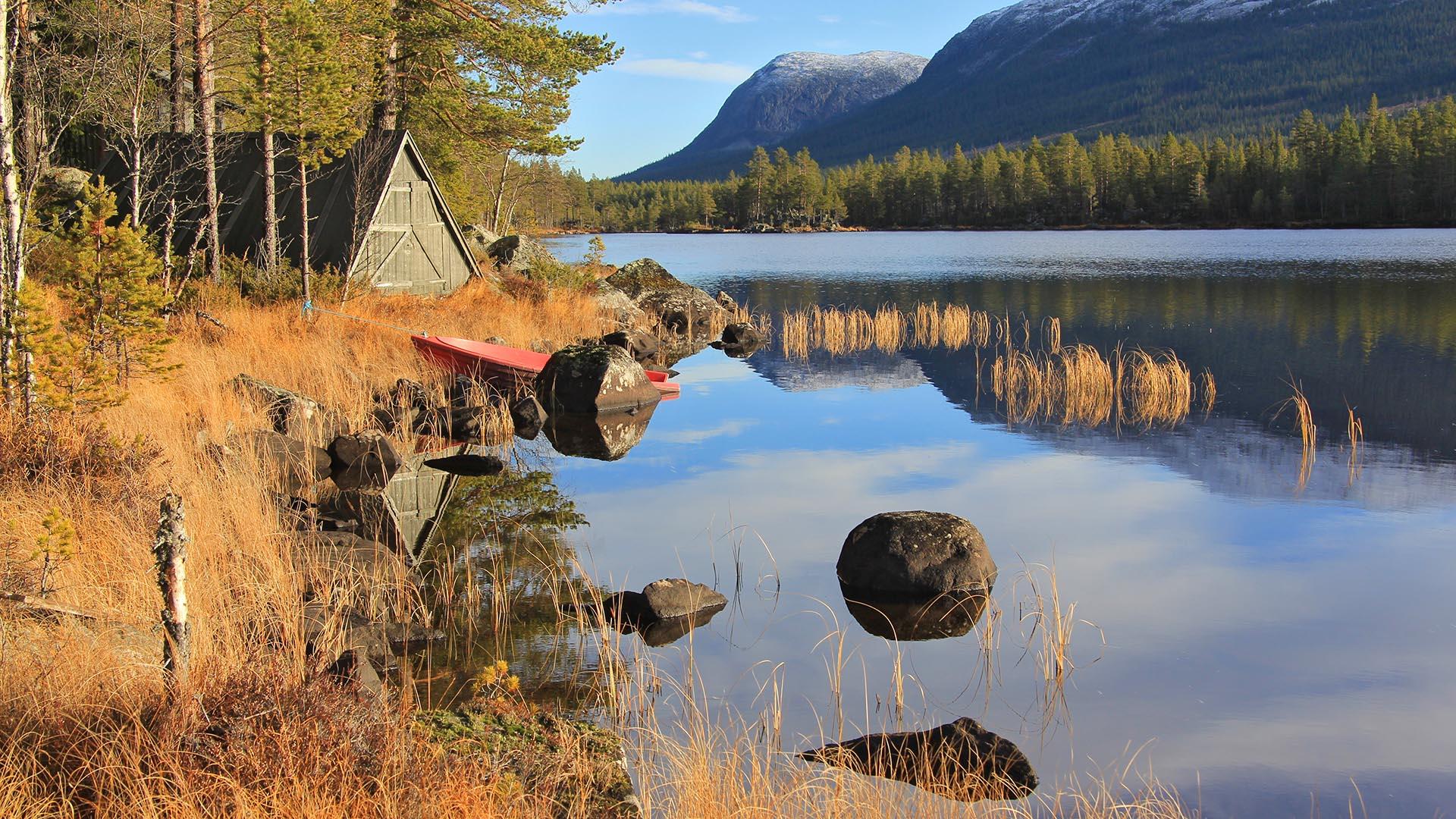 On the lakeshore with tall grass and rocks sits a boat house and a canoe. Mountains and forested hillsides in the background. The pale blue sky is mirrored in the water.