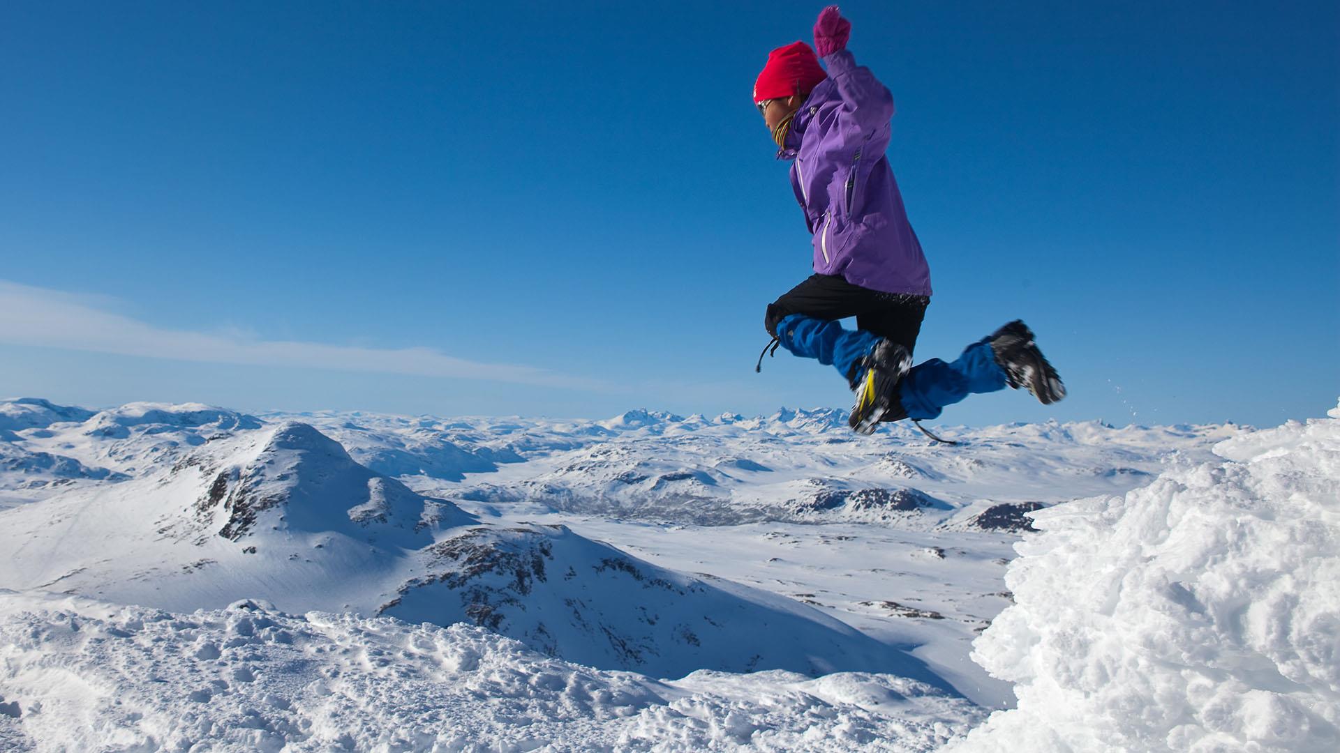A girl in skiing outfit at the summit of a mountain jumps high in the air. Under her feet lies a rugged mountain landscape covered in snow as far as the eye can see. Blue cloudfree sky.