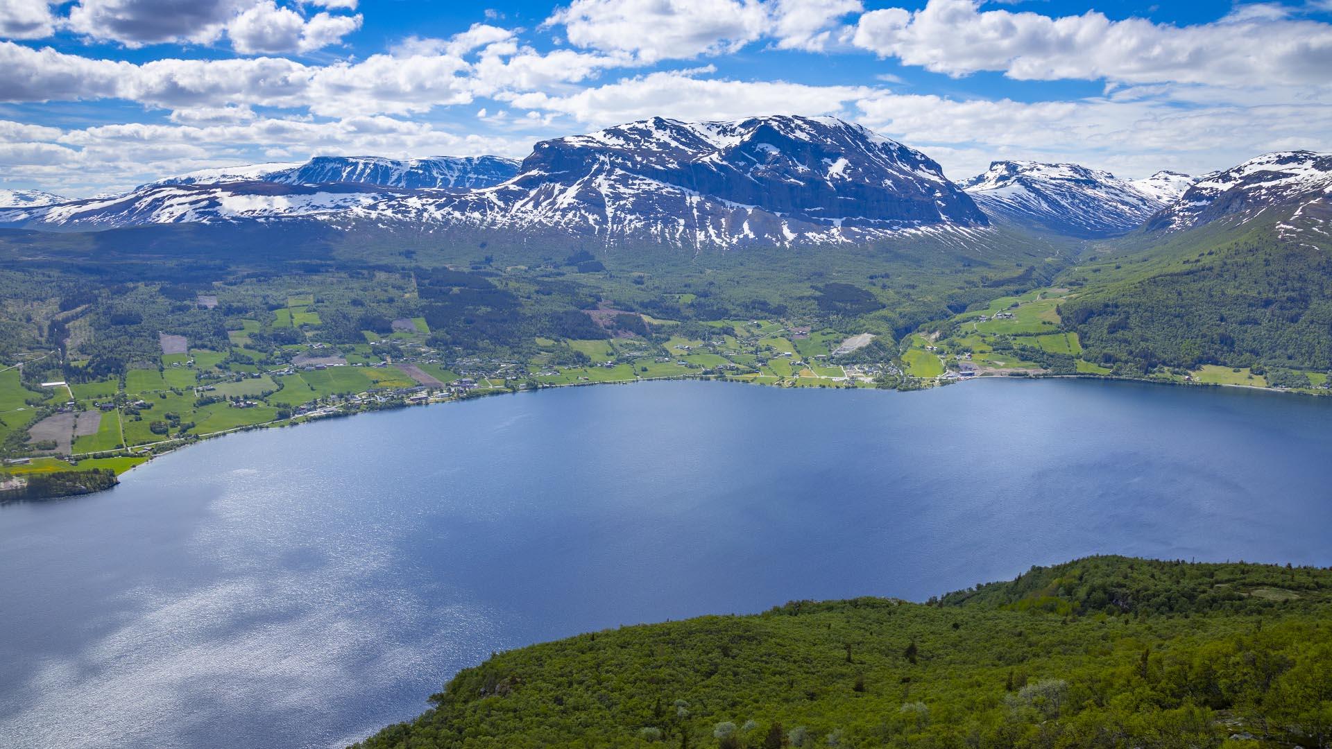 View from a highpoint over a large blue lake. On the other side there are green fields and forets patches on the base of a large mountain massiv with still some snow. Blue sky and white summer clouds.